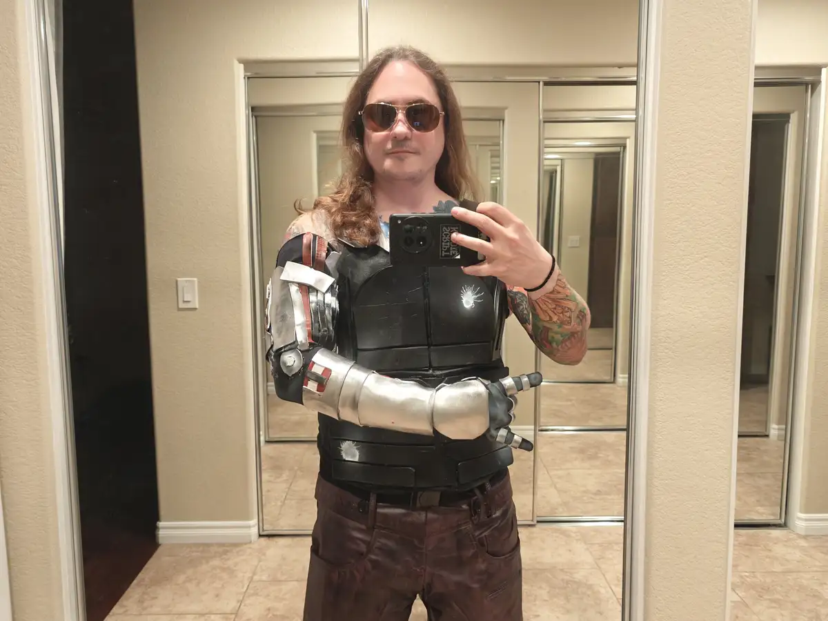 A man with long hair and sunglasses takes a mirror selfie while wearing a detailed black and silver armored vest and a…