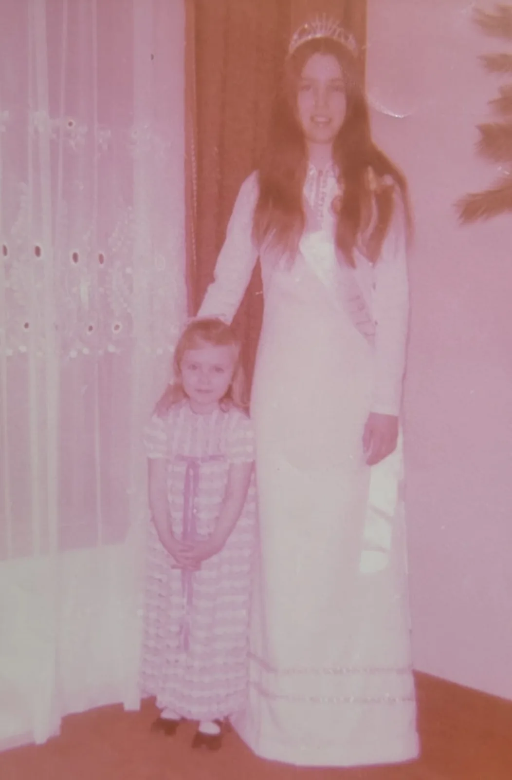 A faded, pink-tinted vintage photo shows a tall young woman in a long white gown and tiara standing beside a small child in…