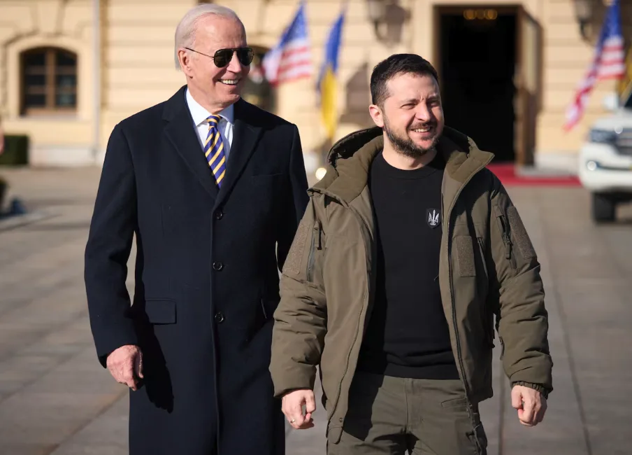 Two men walk side-by-side outdoors in front of a building with flags; the man on the left wears a dark coat and sunglasses…