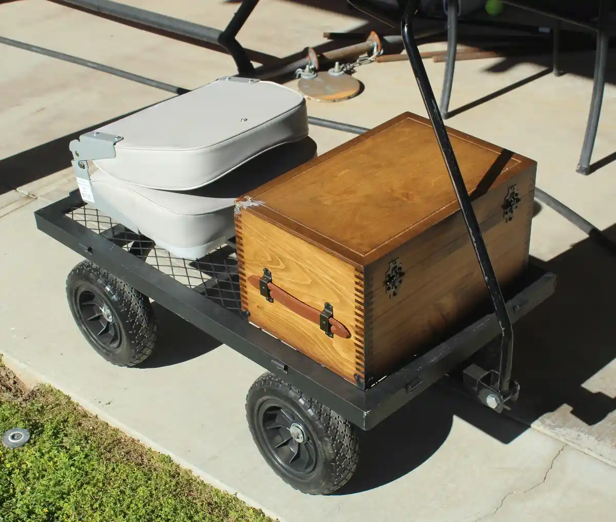 A small black metal cart with two wheels carries a light gray plastic seat and a wooden chest with metal latches and a…
