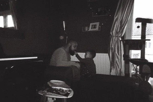 A black-and-white photo shows a man sitting on a couch with a small child beside him. The child is reaching up to touch the…