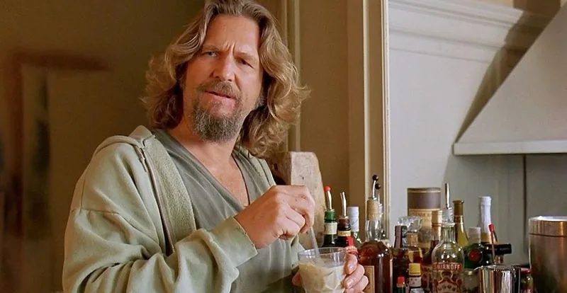 a man with long wavy hair and a beard, wearing a green hoodie, stirs a drink in a glass while standing in front of a bar…