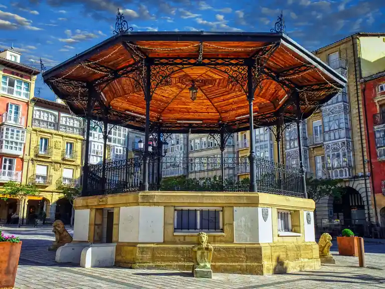A picturesque European plaza features a central ornate gazebo with a wooden roof and black wrought-iron framework…