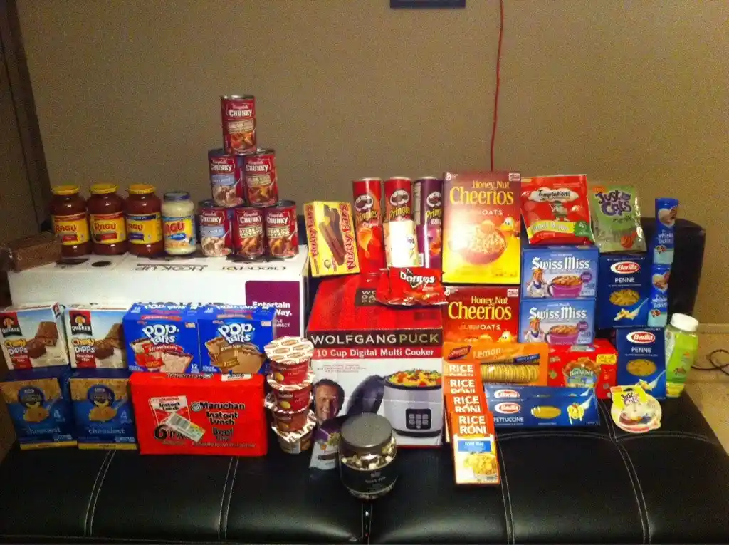 A black leather couch is covered with a massive, chaotic pile of grocery items including pasta, cereal, canned goods…