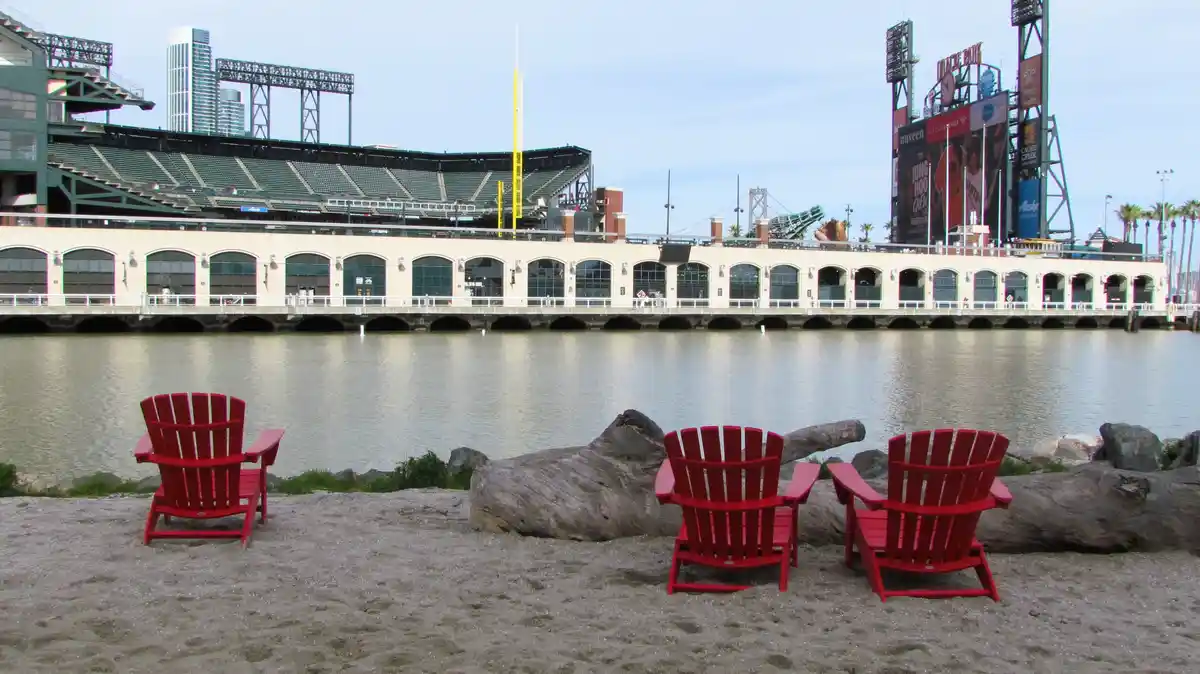 Three bright red Adirondack chairs sit on a sandy beach facing a calm body of water, with the large Oracle Park baseball…
