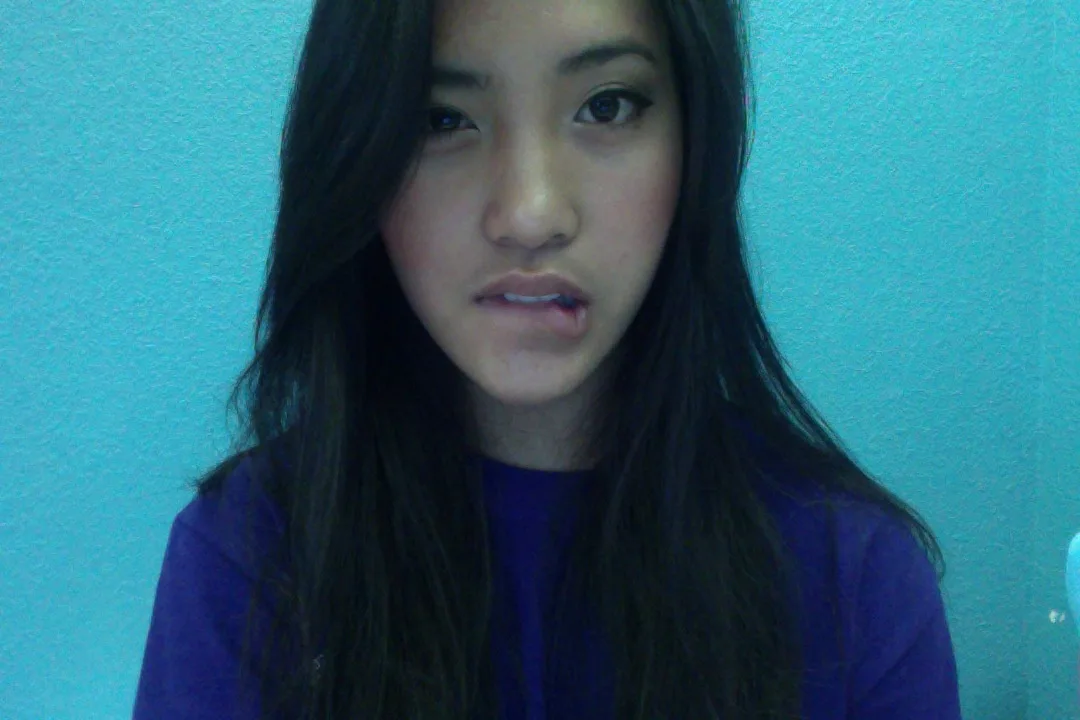 A young woman with long dark hair and a purple shirt stares directly at the camera with a slightly open mouth and a neutral…