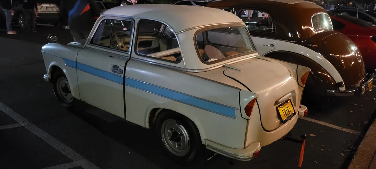 A beige vintage Trabant 600 car with a blue stripe parked at night in a lot, its Virginia license plate reads 'TRABI'. The…