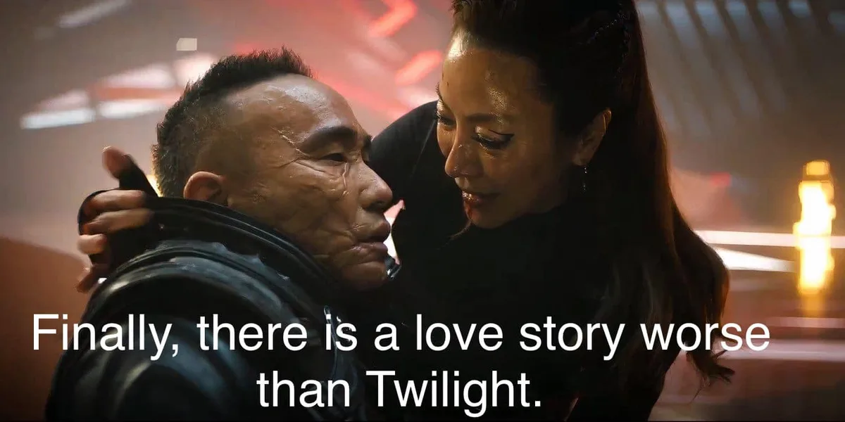 A man with a scarred, sweaty face is embraced by a woman with long dark hair, both looking at each other intimately in a…
