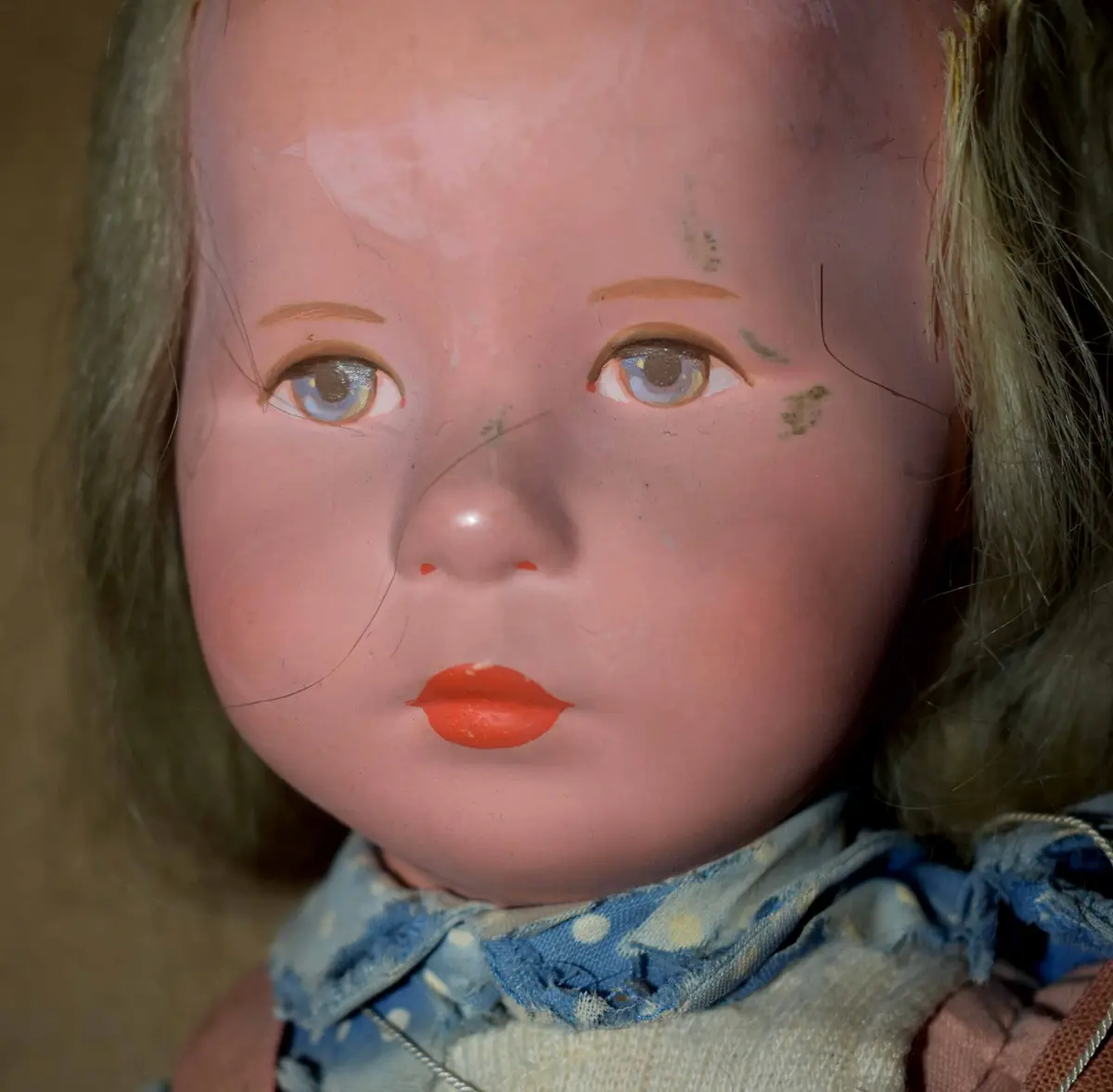 A close-up of a vintage doll with a weathered, pink plastic face, faded blonde hair, and a blue polka-dot collar. The doll…
