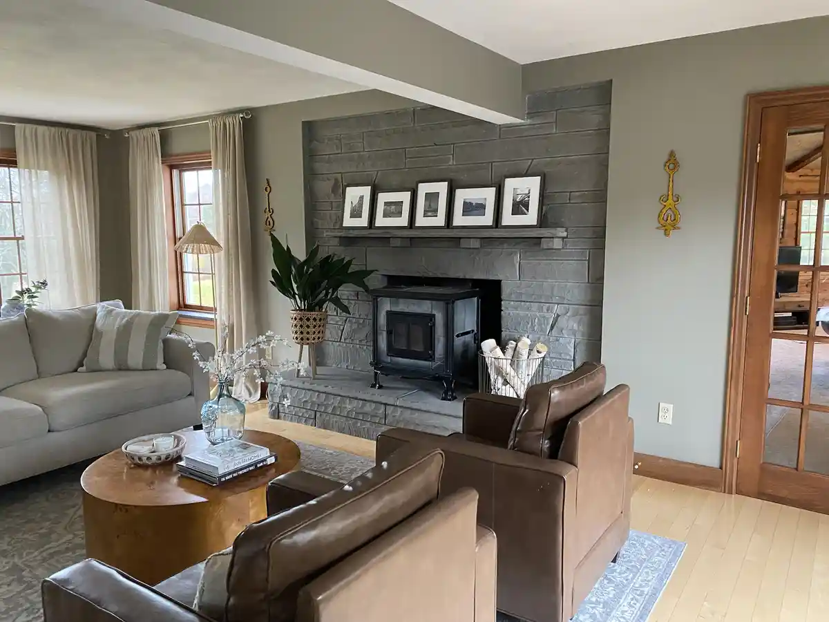 A well-lit, cozy living room with a stone fireplace, leather armchairs, a wooden coffee table, and a gray sofa. The…
