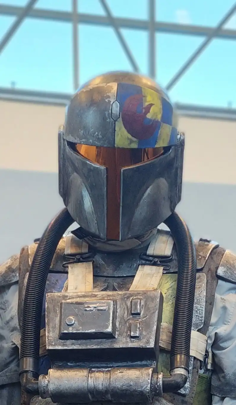 A close-up of a Mandalorian character in a weathered, metallic helmet and armor, with a colorful, painted symbol resembling…