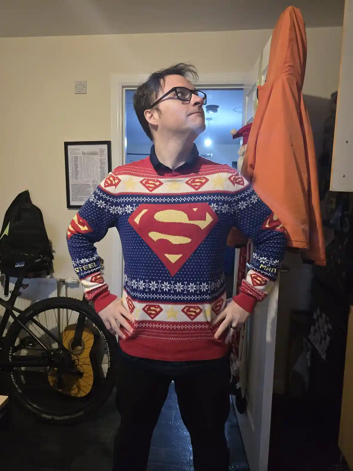 A man wearing a festive Superman-themed Christmas sweater with snowflake patterns and the iconic 'S' logo stands indoors…