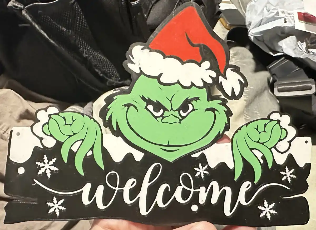 A hand holds a black decorative sign featuring the green, angry face of the Grinch wearing a red and white Santa hat. Below…