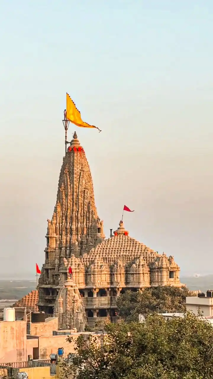 A tall, intricately carved stone temple with a yellow flag at its peak and smaller red flags on other structures, set…