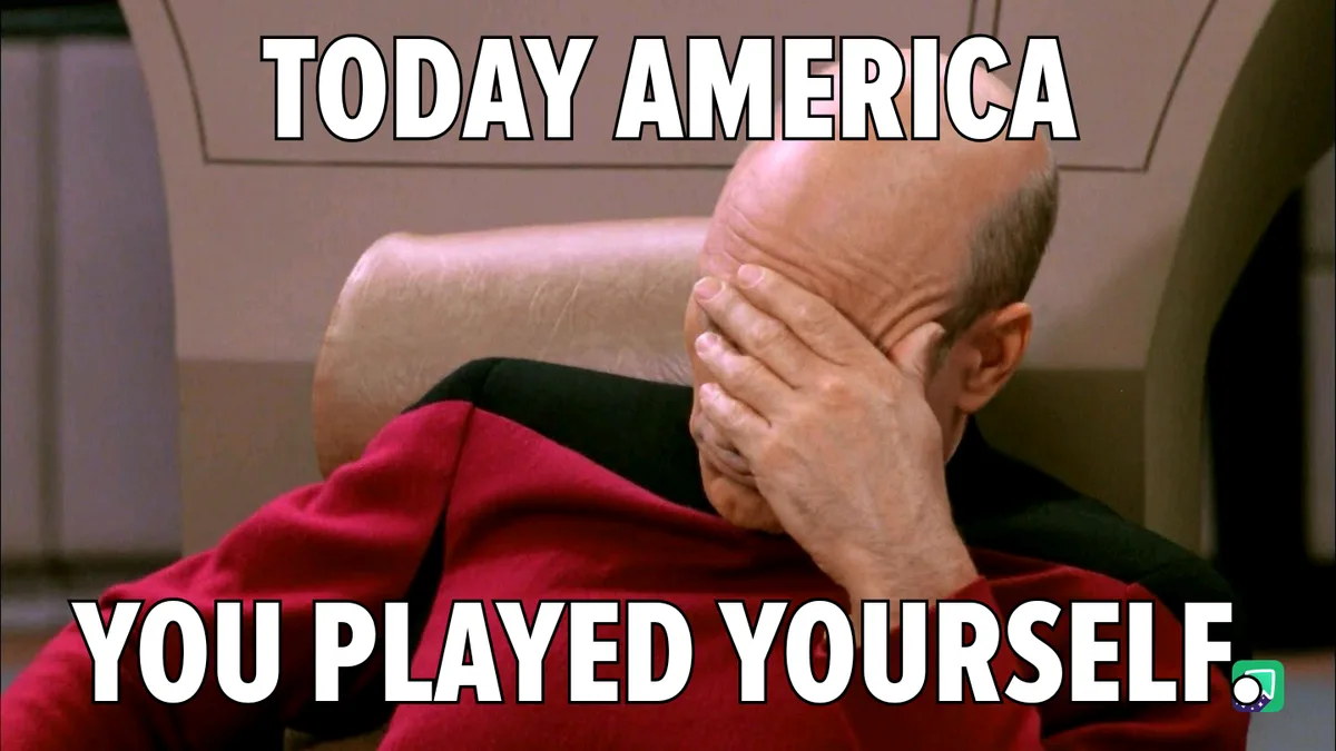 A bald man in a red uniform sits in a chair, covering his face with his hand in a gesture of despair or embarrassment. The…