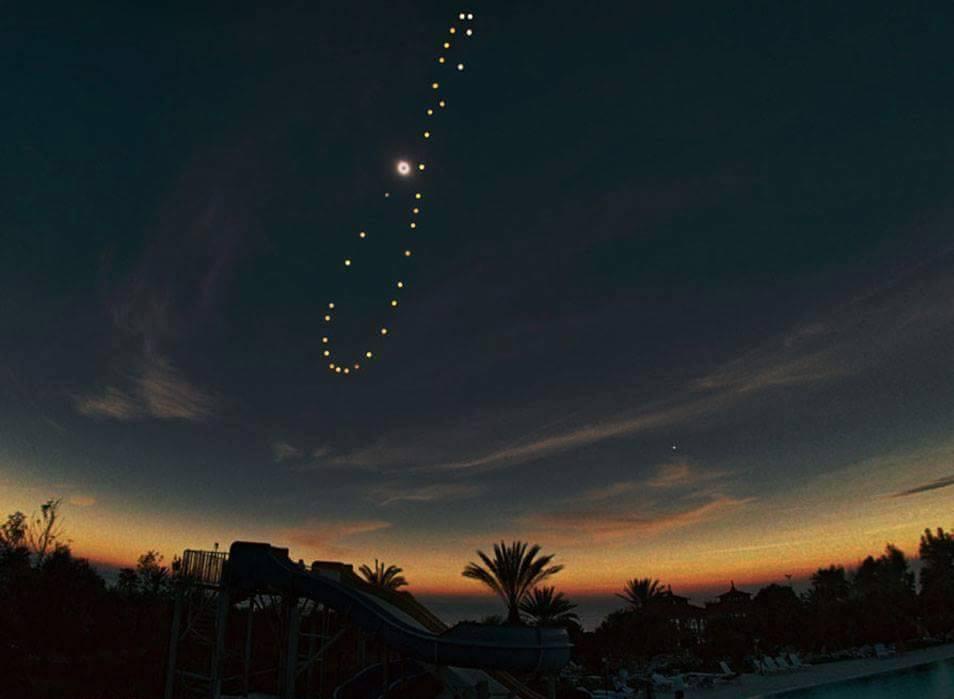 A long-exposure night photo shows a trail of bright dots forming a curved path in the sky above a water park at dusk, with…