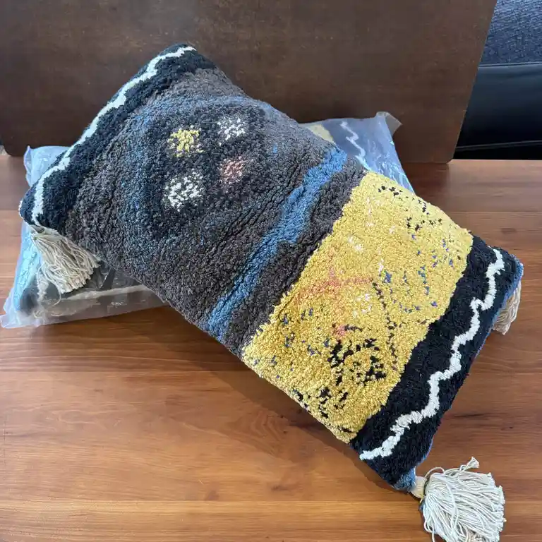 A textured, hand-woven pillow with a dark grey, blue, and yellow pattern and tassels rests on a wooden table, partially…