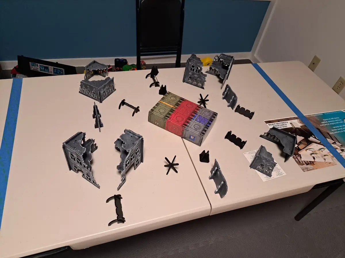 A white table is covered with scattered, unassembled gray plastic game pieces resembling ruined buildings and black…