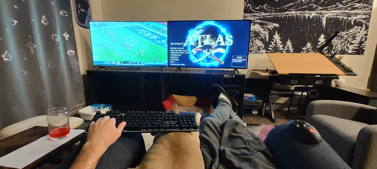 A person is lounging on a couch with their legs up, typing on a keyboard while a dog rests its head on their lap. Two TVs…
