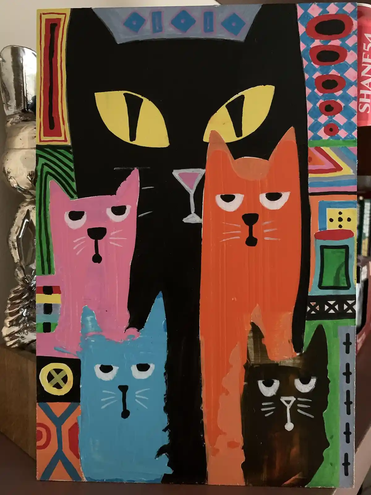 A colorful, stylized painting depicts four cats — a large black cat with yellow eyes, a pink cat, an orange cat, and a blue…