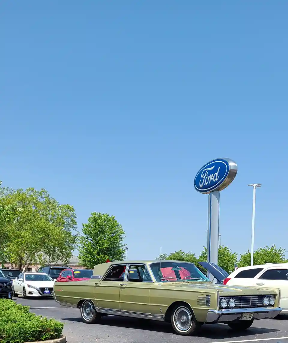 A vintage olive-green Mercury car is parked in a dealership lot under a clear blue sky, with a large Ford dealership sign…