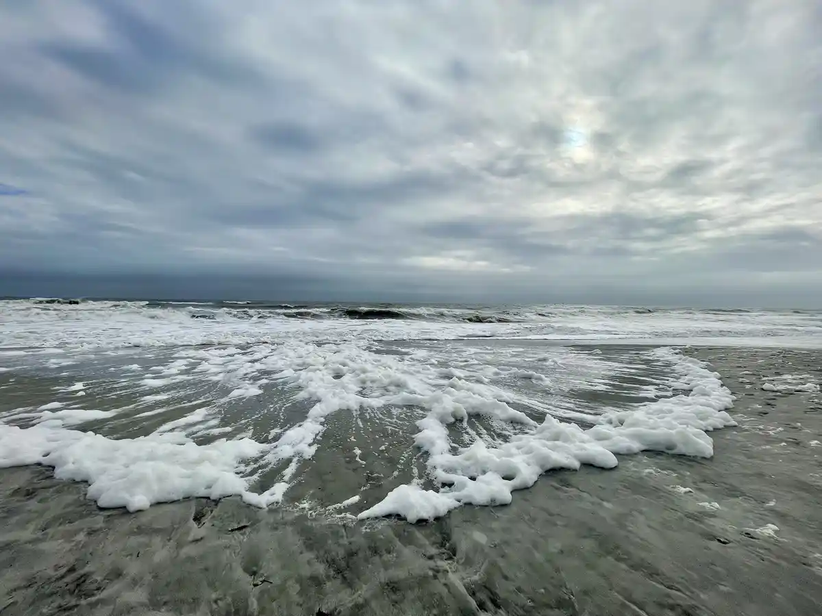 A wide-angle photo of a gray, overcast beach with foamy ocean waves washing onto wet sand. The sky is filled with thick…