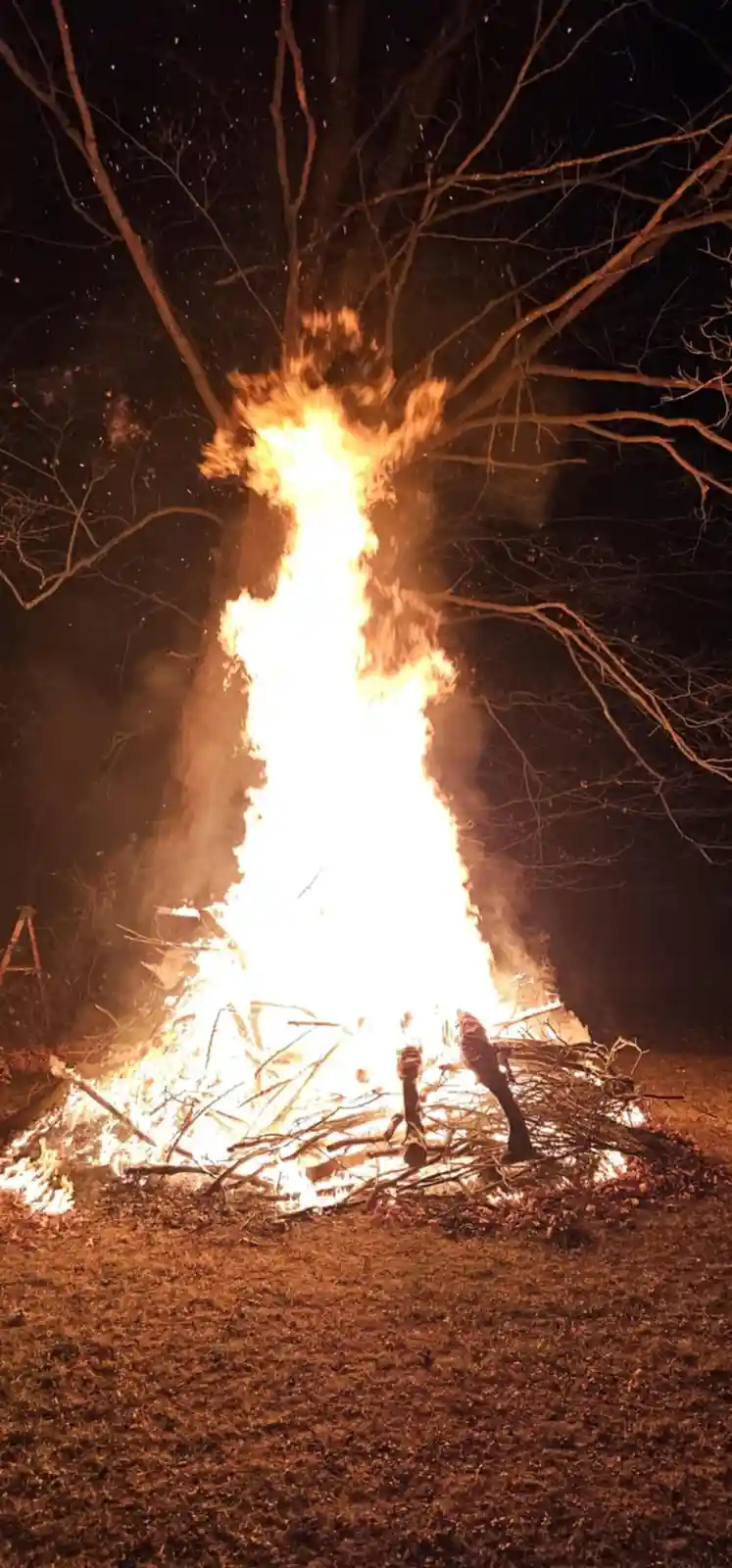 A nighttime photo shows a large bonfire burning intensely, with flames shooting high into the air against a dark sky with…