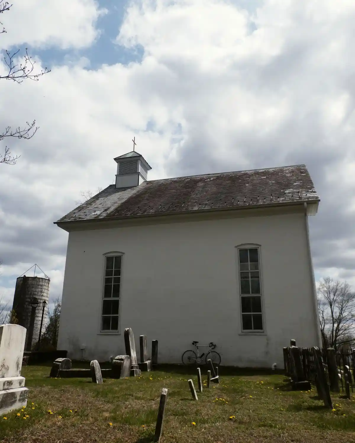 A white church with a cross on its roof stands beside a graveyard with weathered tombstones under a cloudy sky. A silver…