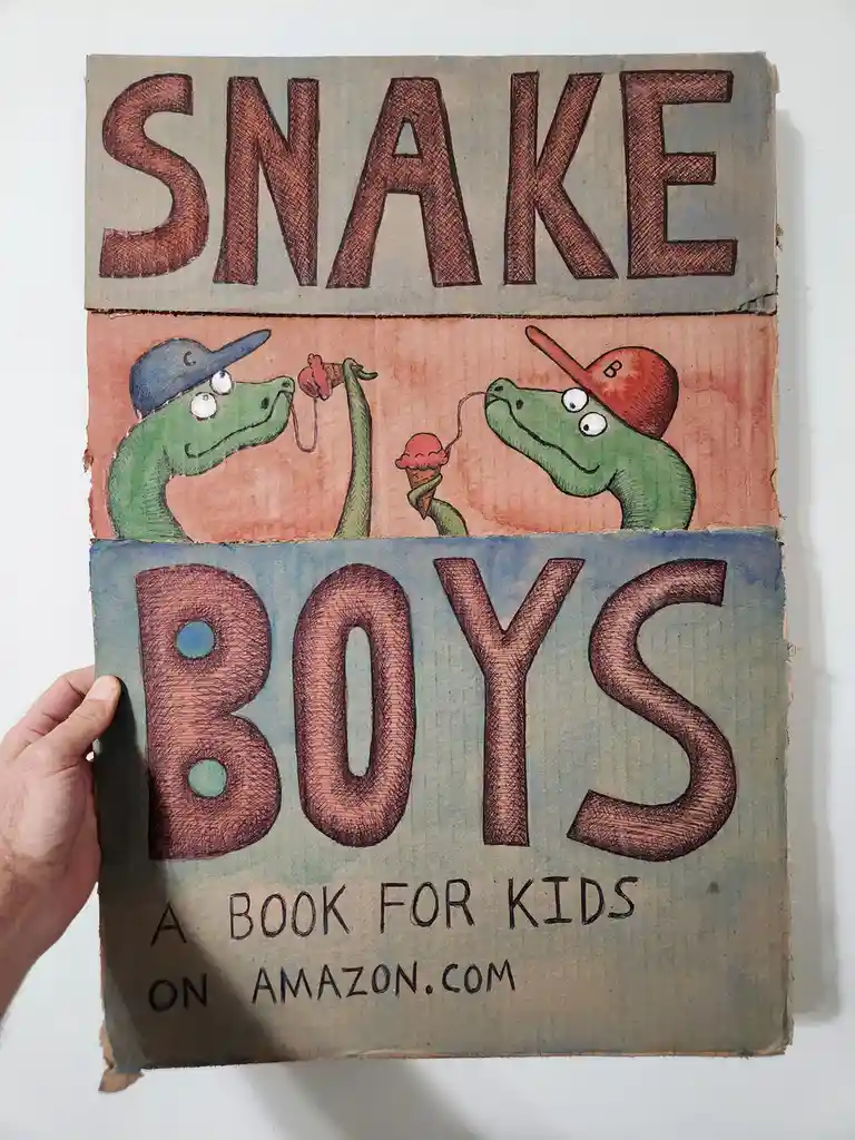 A hand holds a handmade cardboard book cover featuring two cartoon green snakes wearing baseball caps sharing ice cream…