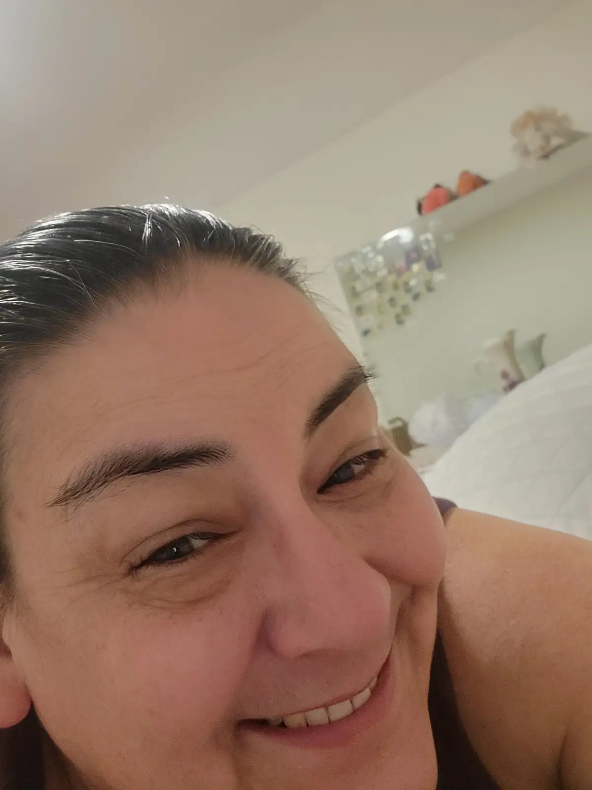 A close-up selfie of a smiling woman with dark hair pulled back, showing crinkled eyes and a wide grin. The background shows…