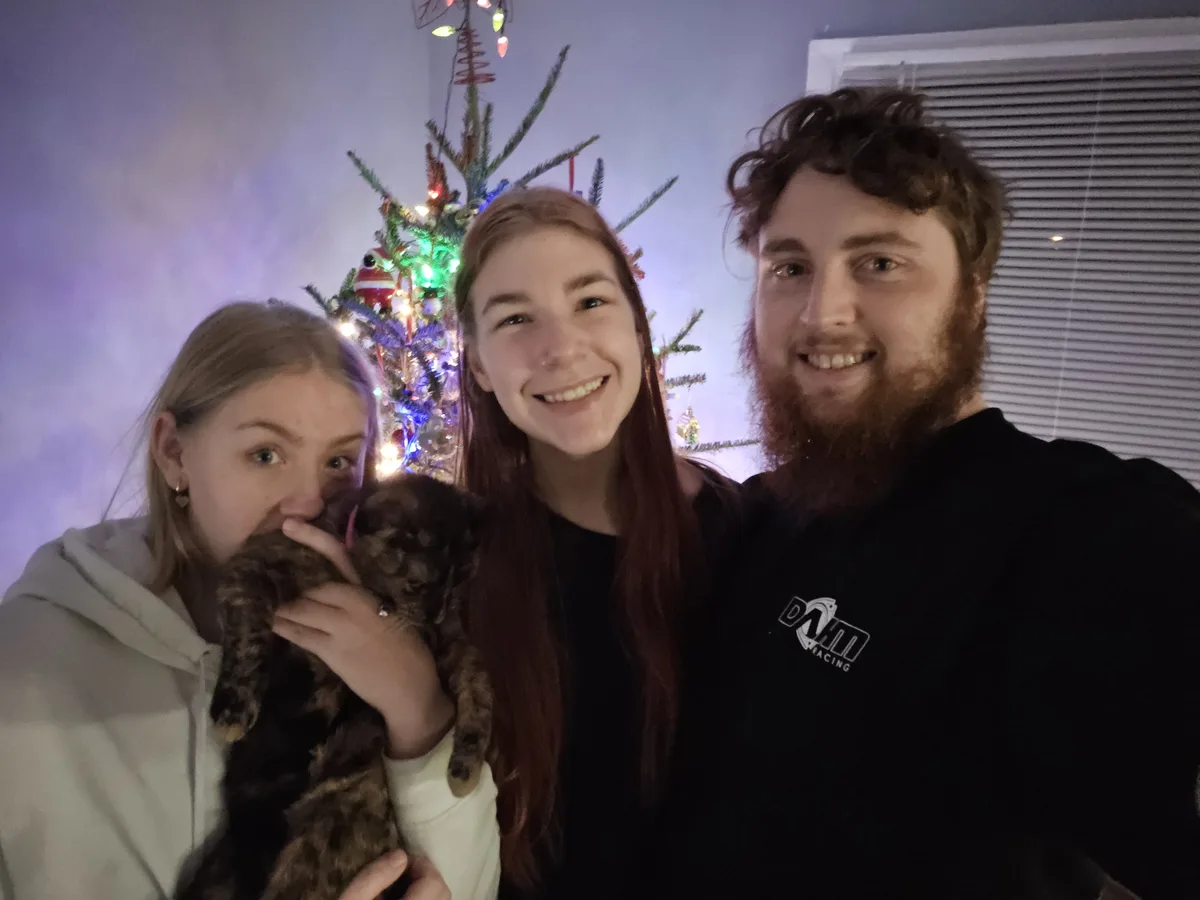 Three people pose for a selfie in front of a decorated Christmas tree; the person on the left holds a small brown cat close…