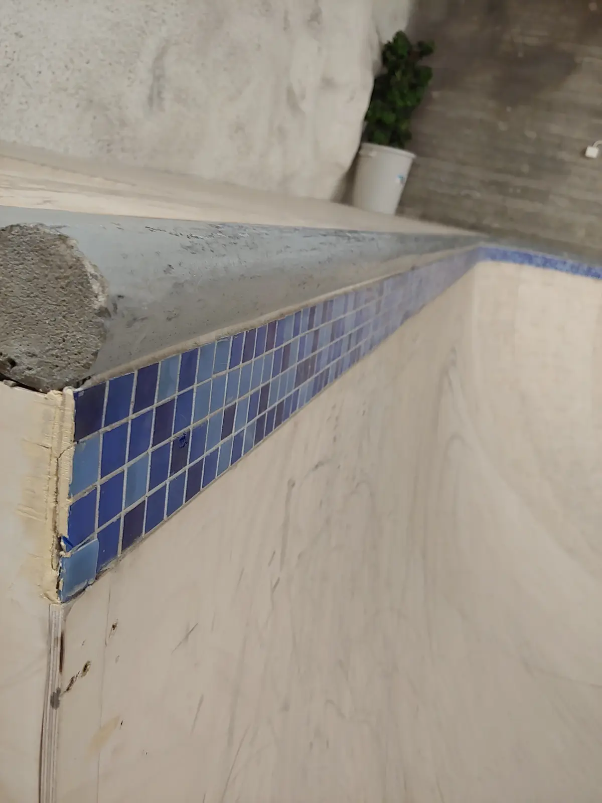 A close-up photo of a partially tiled pool edge with blue mosaic tiles, showing a rough concrete coping and unfinished…