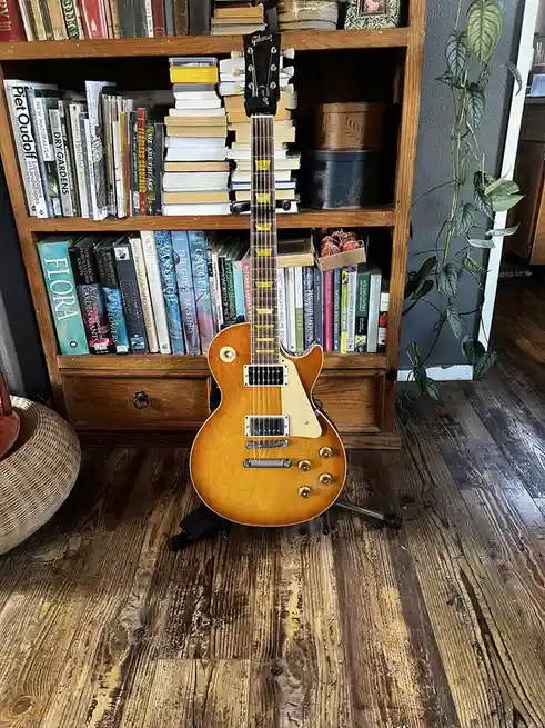 A Gibson Les Paul electric guitar stands upright on a black guitar stand on a dark wood floor, positioned in front of a…