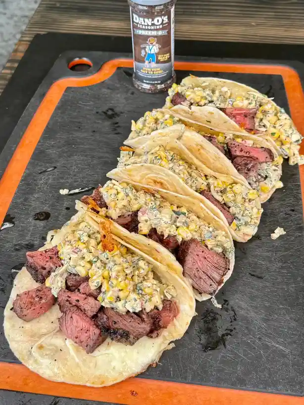 A row of five tacos filled with sliced steak and creamy corn salsa sits on a black cutting board with an orange border. A…