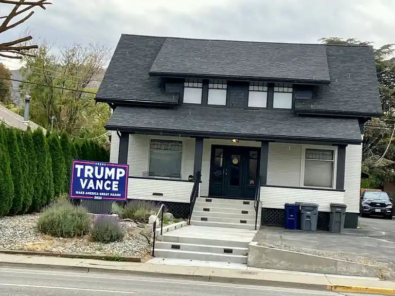 A residential house with white siding and dark trim has a large blue political yard sign in front that reads 'TRUMP VANCE…
