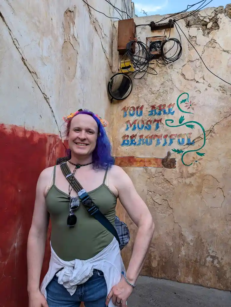 A person with purple hair and a colorful headband smiles at the camera while standing in front of a weathered wall. The wall…