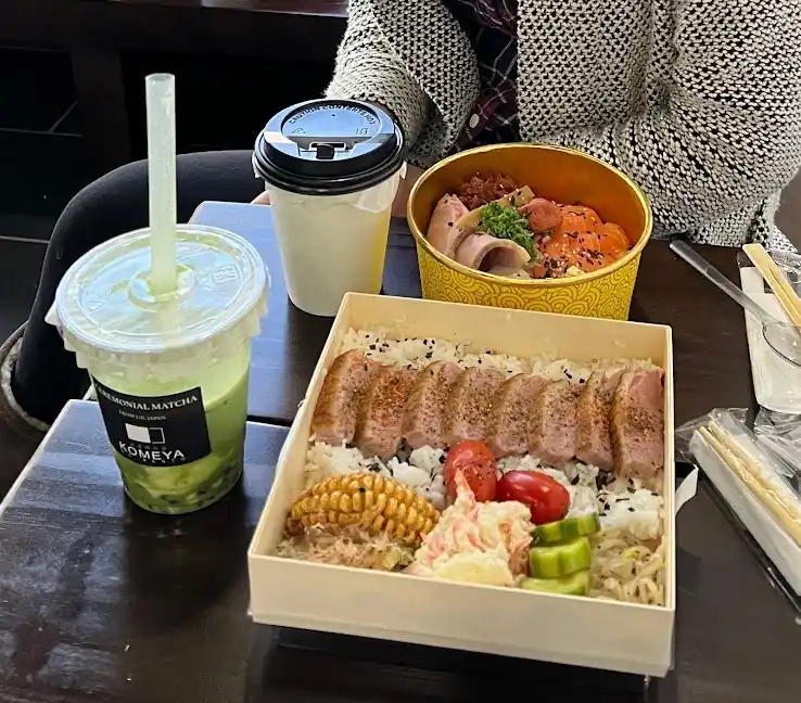 A table displays two Japanese meals and drinks: a bento box with rice, sliced meat, corn, tomatoes, and pickles, and a…