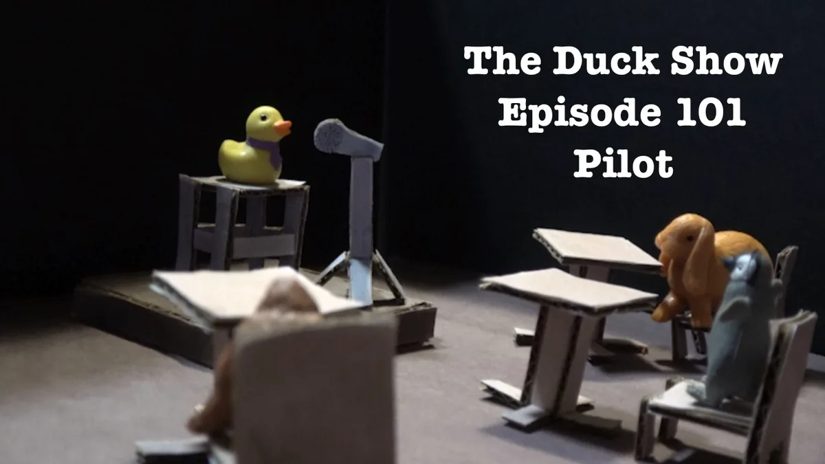 A stop-motion scene shows a yellow rubber duck wearing a purple scarf standing on a cardboard podium with a microphone…