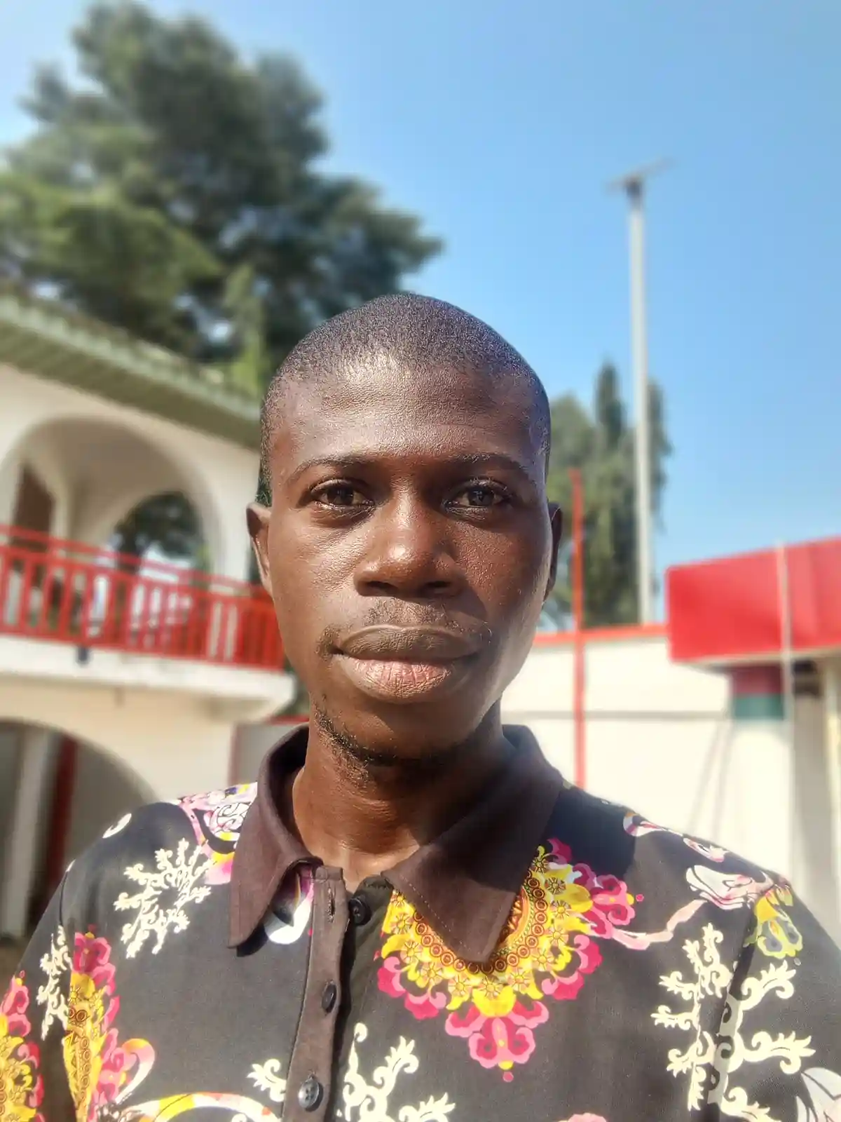 A man with a shaved head and a neutral expression stares directly at the camera, wearing a dark shirt with colorful floral…