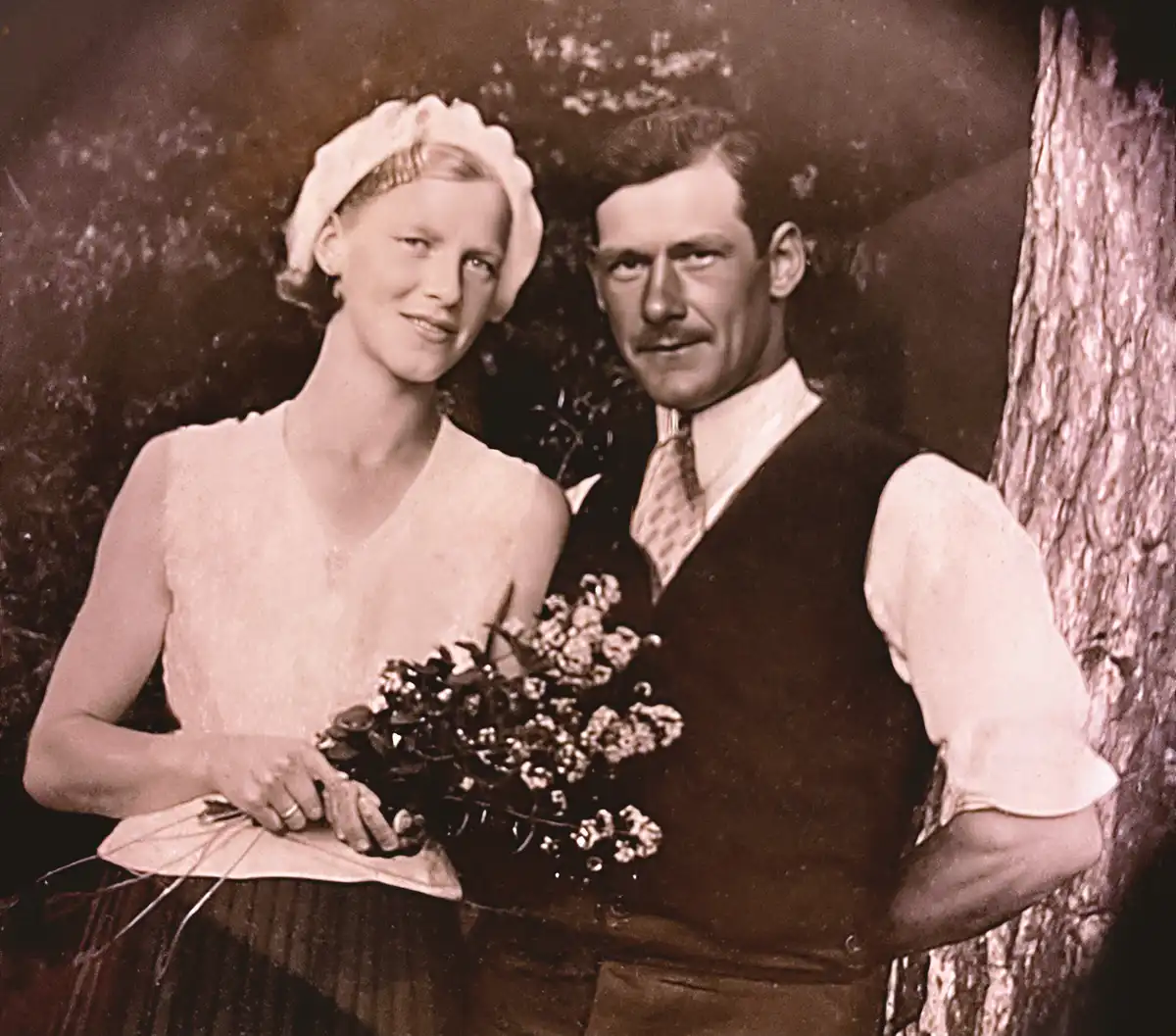 A vintage sepia-toned wedding photo shows a smiling bride in a white dress and hat holding a bouquet, standing beside a…