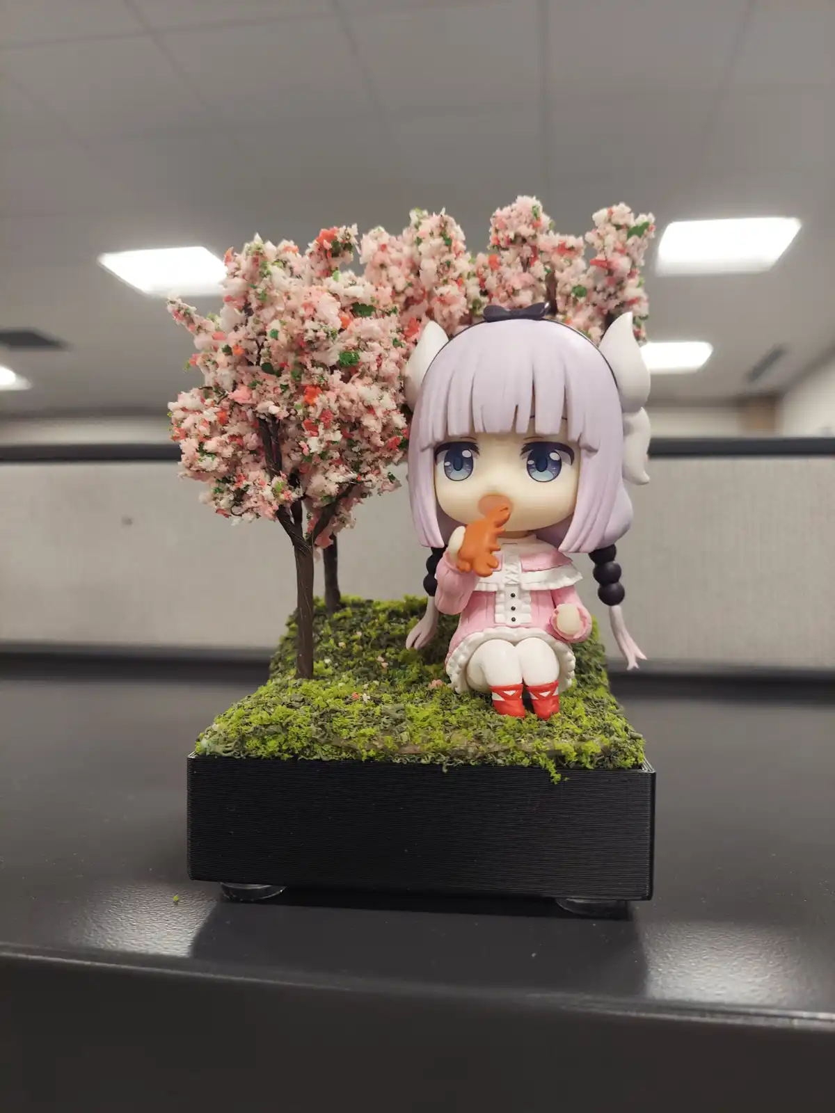 A chibi-style anime figurine with light purple hair, white horns, and blue eyes sits on a mossy base next to miniature…