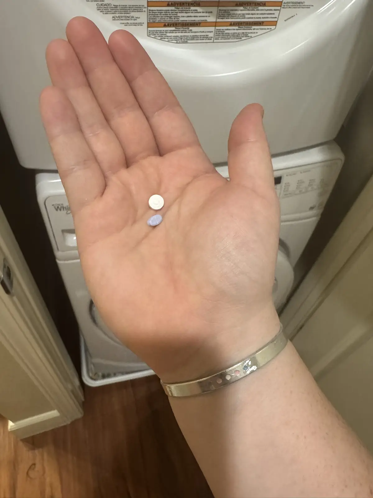 A person's open palm displays two small pills, one white and one light purple, held in front of a white stacked washer and…