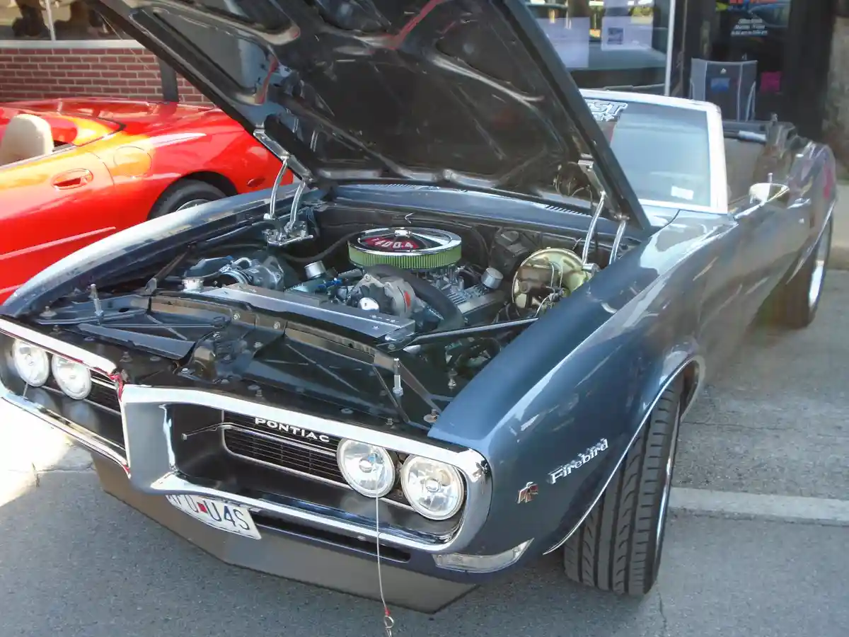 A blue Pontiac Firebird convertible with its hood open, revealing a large 400 cubic inch engine. The car is parked outdoors…