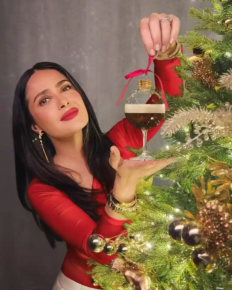 A woman in a red top stands beside a decorated Christmas tree, holding a glass ornament shaped like a wine glass filled with…