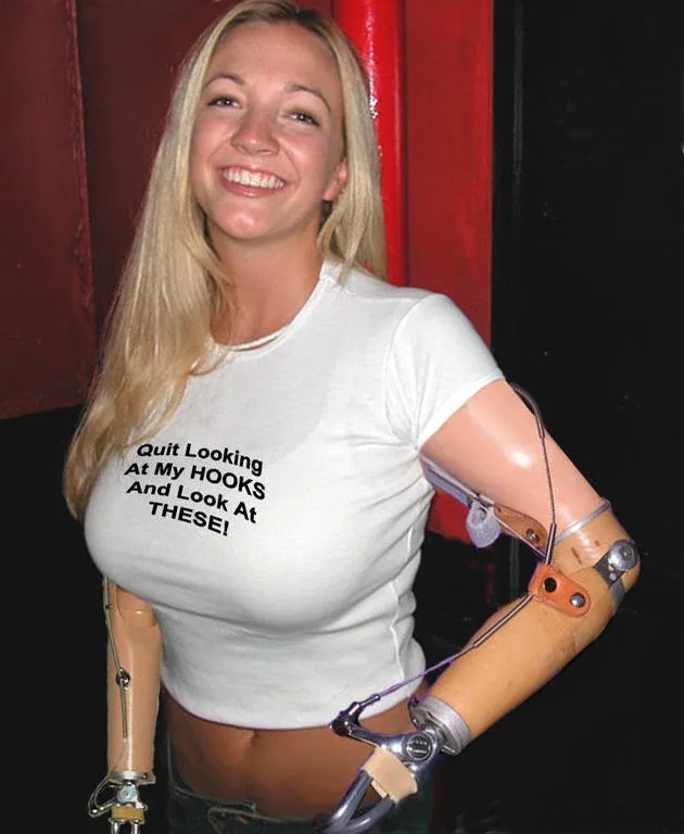 A smiling blonde woman with prosthetic arms wears a white t-shirt with the text 'Quit Looking At My HOOKS And Look At…