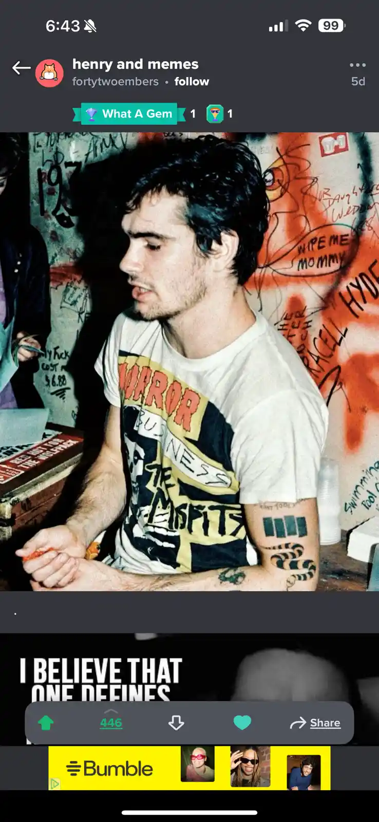 A man with dark hair and tattoos on his arms sits in front of a graffiti-covered wall, wearing a graphic t-shirt. The…