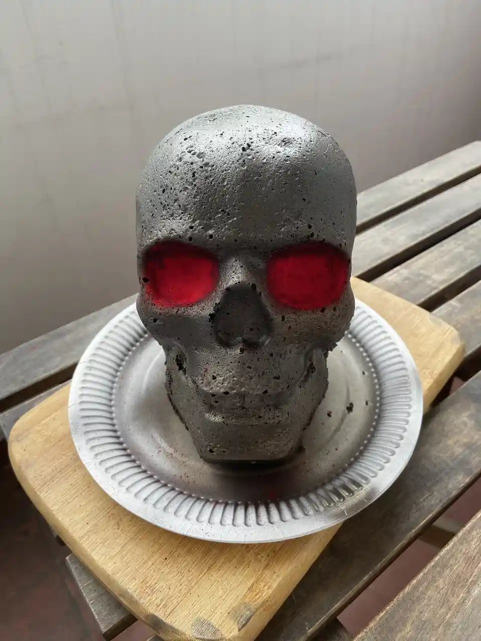 A grey, textured skull-shaped cake with bright red glowing eyes sits on a white paper plate, which rests on a wooden cutting…