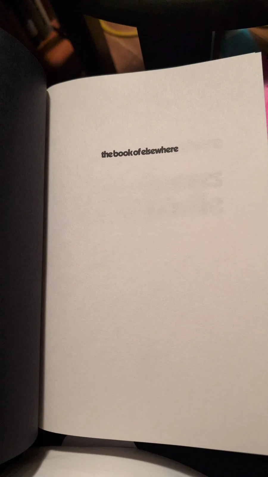 An open book displays a blank white page with the text 'the book of elsewhere' centered in a simple black font. The…