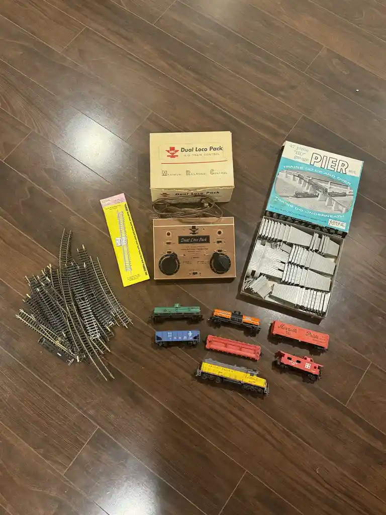 An overhead shot displays a collection of vintage HO gauge model train components laid out on a dark wood floor, including a…