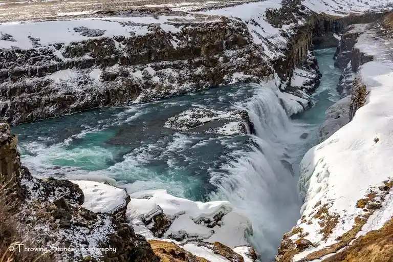 A wide-angle photograph of a powerful, snow-dusted waterfall cascading through a deep canyon with icy cliffs and turquoise…